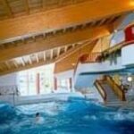 Hallenbadbereich mit Holzdecke, großen Fenstern, Treppen und Menschen, die im Wasser schwimmen.