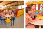 Vier kleine Kinder in Badeanzügen sitzen um einen kleinen runden Tisch mit Getränken und Snacks in einem farbenfrohen Indoor-Spielbereich in Form eines Bootes.