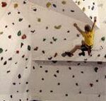 Eine Person in einem gelben Hemd klettert an einer Indoor-Kletterwand mit bunten Griffen.