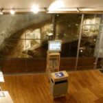 Eine moderne Museumsausstellung mit Holzfußboden, Ausstellungsständern, Informationsmaterial und einer Glaswand, hinter der sich eine archäologische Ausgrabungsstätte befindet.