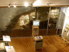 Eine moderne Museumsausstellung mit Holzfußboden, Ausstellungsständern, Informationsmaterial und einer Glaswand, hinter der sich eine archäologische Ausgrabungsstätte befindet.