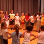 Eine Gruppe von Kindern steht in einem hell erleuchteten Raum im Kreis und hält sich an den Händen, während ein Erwachsener die Aktivität leitet.