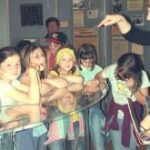 Eine Gruppe von Kindern steht um eine Vitrine herum, während ein Erwachsener auf sie zeigt und spricht, wahrscheinlich um eine Erklärung oder eine Führung in einem Museum oder einer Bildungseinrichtung zu geben.