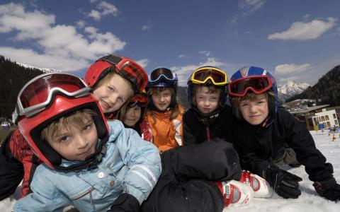 Sechs Kinder in Winterkleidung und Skihelmen posieren gemeinsam auf einer verschneiten Piste unter einem teilweise bewölkten Himmel mit Bergen im Hintergrund.