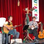 Zwei Musiker spielen auf der Bühne mit Gitarren, einem Akkordeon und Notenständern vor einem roten Vorhang und einem bunten "UTAKIRO"-Banner im Hintergrund.