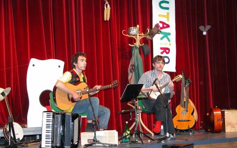 Zwei Musiker spielen auf der Bühne mit Gitarren, einem Akkordeon und Notenständern vor einem roten Vorhang und einem bunten "UTAKIRO"-Banner im Hintergrund.