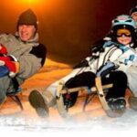 Zwei Erwachsene und zwei Kinder fahren nachts in Winterkleidung und mit Helmen auf Holzschlitten einen verschneiten Hügel hinunter.