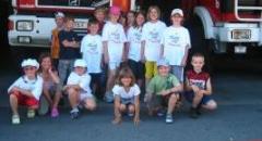 Eine Gruppe von Kindern posiert vor einem Feuerwehrauto, einige stehen, andere hocken, alle tragen Freizeitkleidung und lächeln.