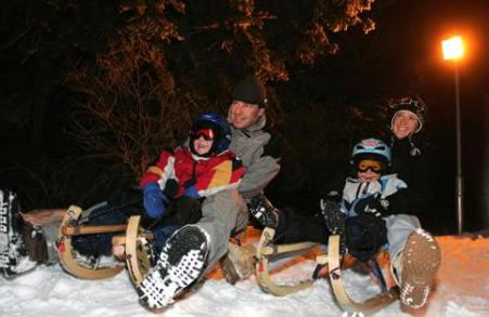 Vier Personen in Winterkleidung, die nachts auf Schlitten im Schnee sitzen und lächeln, mit einer Straßenlaterne und Bäumen im Hintergrund.