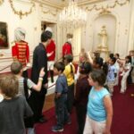 Eine Gruppe von Kindern betrachtet eine Ausstellung von Soldatenpuppen in historischen Uniformen in einem geschmückten, von Kronleuchtern beleuchteten Raum.