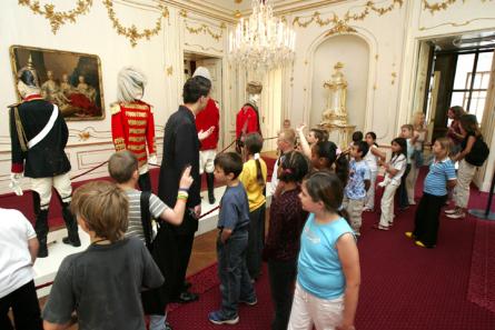 Eine Gruppe von Kindern betrachtet eine Ausstellung von Soldatenpuppen in historischen Uniformen in einem geschmückten, von Kronleuchtern beleuchteten Raum.