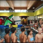 Kinder in Badeanzügen versammeln sich um ein Hallenbad mit aufblasbaren Spielgeräten; im Hintergrund sind Erwachsene und weitere Kinder zu sehen.