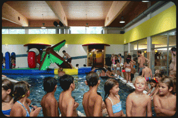 Kinder in Badeanzügen versammeln sich um ein Hallenbad mit aufblasbaren Spielgeräten; im Hintergrund sind Erwachsene und weitere Kinder zu sehen.
