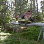 Zwei Personen fahren auf einer einspurigen Alpenachterbahn durch ein bewaldetes Gebiet, umgeben von Bäumen und Grünflächen.