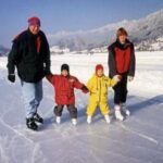 Eine vierköpfige Familie in Winterkleidung und Schlittschuhen hält sich an den Händen, während sie im Freien auf einer gefrorenen Fläche mit verschneiten Bergen im Hintergrund Schlittschuh läuft.