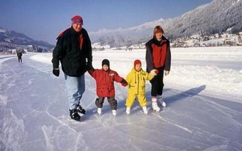 Eine vierköpfige Familie in Winterkleidung und Schlittschuhen hält sich an den Händen, während sie im Freien auf einer gefrorenen Fläche mit verschneiten Bergen im Hintergrund Schlittschuh läuft.