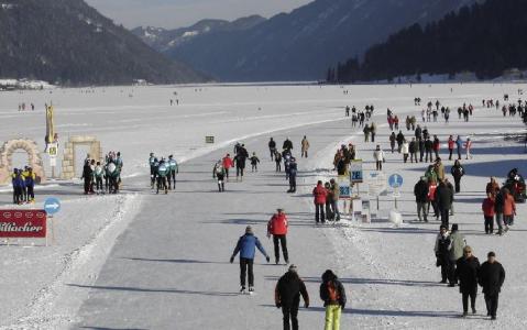 Auf einem großen zugefrorenen See, der von schneebedeckten Bergen umgeben ist, gehen die Menschen spazieren und laufen Schlittschuh, und entlang des Eises sind Gruppen versammelt und Schilder aufgestellt.