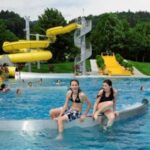 Zwei Mädchen sitzen am Rande eines öffentlichen Freibads mit einer gelben Wasserrutsche und anderen Badegästen im Hintergrund, umgeben von Bäumen und Grünflächen.