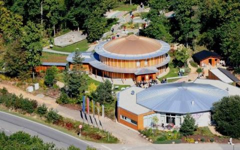 Luftaufnahme eines kreisförmigen Gebäudekomplexes aus Holz, umgeben von Bäumen und Wegen, mit mehreren Personen, die herumlaufen, und geparkten Autos in der Nähe des Eingangs.