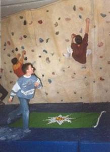 Drei Kinder klettern an einer Indoor-Kletterwand über blaue Matten, auf dem Boden liegt eine grüne Matte mit einer weißen Blume.