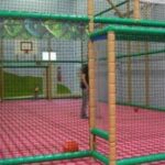 Spielbereich im Innenbereich mit gepolstertem Boden, Netzeinfassung, verstreuten Bällen und einem Basketballkorb. Ein Kind und ein Erwachsener befinden sich in der Spielzone.