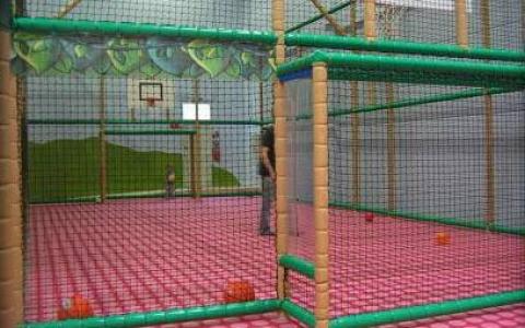 Spielbereich im Innenbereich mit gepolstertem Boden, Netzeinfassung, verstreuten Bällen und einem Basketballkorb. Ein Kind und ein Erwachsener befinden sich in der Spielzone.