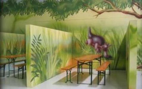 Holztische und -bänke stehen zwischen Wänden, die mit einem Dschungelthema bemalt sind, darunter Grünpflanzen und die Abbildung eines Dinosauriers.