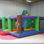 Aufblasbares Indoor-Spielgerät mit Kletterwand und bunten Säulen auf einem rot-weiß karierten Boden in einem geräumigen Raum.