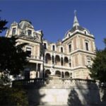 Großes historisches Herrenhaus mit verschnörkelter Architektur, Ziegel- und Steinfassade, zahlreichen Fenstern, Balkonen, Türmen und einem Spitzdach, umgeben von Bäumen unter einem klaren blauen Himmel.
