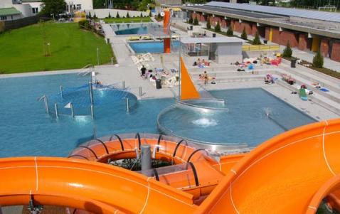 Blick auf ein Freibad mit mehreren Schwimmbecken, Lounge-Bereichen und leuchtend orangefarbenen Wasserrutschen. Menschen nehmen ein Sonnenbad und schwimmen.