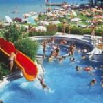 Menschen schwimmen und spielen auf Wasserrutschen in einem überfüllten Freibad neben einer Grünfläche mit Sonnenschirmen und Sonnenanbetern, mit einem See im Hintergrund.