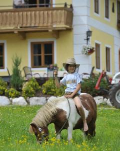 Ein Kind mit Hut sitzt barfuß auf einem kleinen Pony, das auf einer Wiese mit einem gelben Haus und Blumen im Hintergrund weidet.