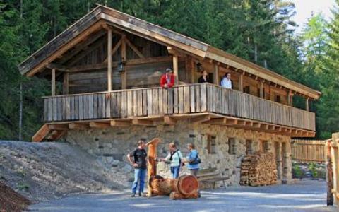 Ein rustikales Holzhaus mit Steinsockel ist von Bäumen umgeben; mehrere Menschen stehen auf dem Balkon und bei den gestapelten Holzscheiten im Freien.