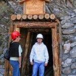 Zwei Personen mit Schutzhelmen stehen am Eingang eines in einen felsigen Abhang gebauten Minenschachts mit Holzrahmen.