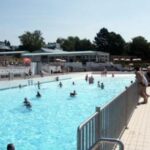 Freibad mit Menschen im Wasser und anderen, die am Beckenrand stehen; an einem sonnigen Tag sind im Hintergrund Gebäude und Bäume zu sehen.