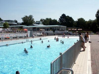 Freibad mit Menschen im Wasser und anderen, die am Beckenrand stehen; an einem sonnigen Tag sind im Hintergrund Gebäude und Bäume zu sehen.