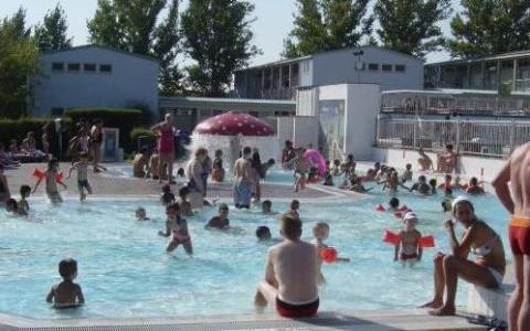 Kinder und Erwachsene schwimmen und spielen in einem überfüllten Freibad an einem sonnigen Tag, mit Gebäuden und Bäumen im Hintergrund.