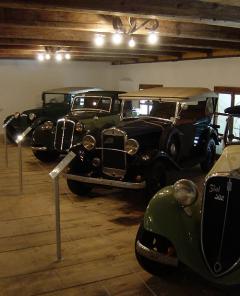 Vier Oldtimer sind in einem Innenraum auf einem Holzboden unter Deckenbeleuchtung ausgestellt, jeder hinter einem Informationsständer aus Metall.