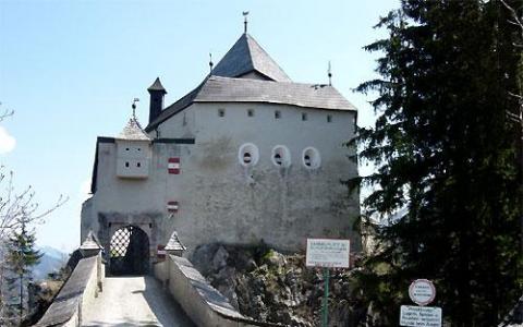 Ein steinernes Schloss mit schrägem Dach, runden Fenstern und Fahnen steht am Ende einer Brücke, umgeben von Bäumen und Schildern am Eingang.