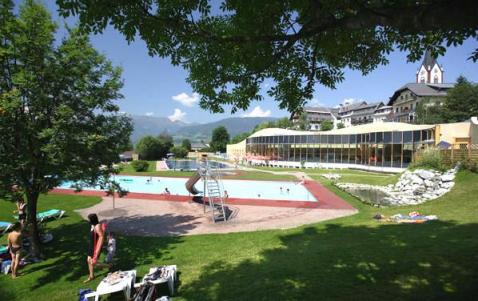 Freibad mit Rutsche, umgeben von Gras, Bäumen und ein paar Menschen; im Hintergrund sind Gebäude und Berge zu sehen.