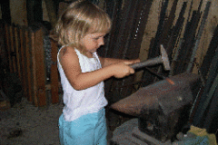 Ein junges Kind in einem weißen Tank-Top und blauen Shorts benutzt einen Hammer auf einem Amboss in einer Werkstattumgebung.