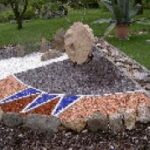 Ein kleiner, mit Steinen eingefasster Steingarten mit einem großen aufrechten Felsen und buntem Kies, der in geometrischen Mustern angeordnet ist. Im Hintergrund sind grünes Gras und Pflanzen zu sehen.