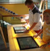 Zwei Personen verwenden Touchscreen-Displays an einem Holztisch in einem hell erleuchteten Innenraum.