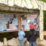 Zwei Kinder stehen vor den Informationstafeln im Freien und lesen die Schilder unter einem Unterstand aus Stein und Holz.