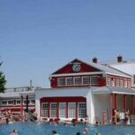 Menschen schwimmen in einem Freibad vor einem rot-weißen Gebäude mit großen Fenstern und einer Uhr an der oberen Fassade, unter einem klaren blauen Himmel.