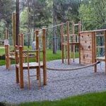Spielplatz aus Holz mit Kletterelementen, einer Seilbrücke und einem Kiesboden, umgeben von Bäumen und grünem Gras.