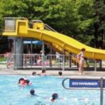 Menschen schwimmen in einem Freibad in der Nähe einer gelben Wasserrutsche; eine Frau steht am Eingang der Rutsche. Im Hintergrund sind Bäume und ein "Schwimmer"-Schild zu sehen.