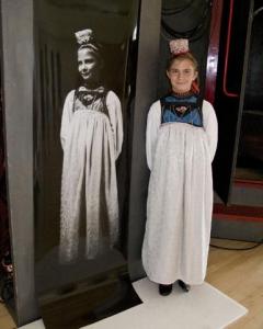 Ein Mädchen in traditioneller Kleidung steht neben einem großen Schwarz-Weiß-Foto eines anderen Mädchens in ähnlicher Kleidung.