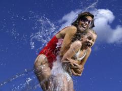Zwei Personen in Badeanzügen lachen und planschen im Wasser vor einem strahlend blauen Himmel.
