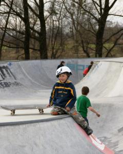 Ein kleines Kind mit einem Helm sitzt mit einem Skateboard am Rand einer Skateparkrampe, während ein anderes Kind in einem grünen T-Shirt in der Nähe steht.
